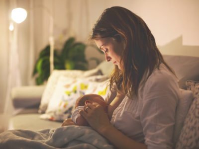 Young beautiful mother, breastfeeding her newborn baby boy at night, dim light. Mom breastfeeding infant