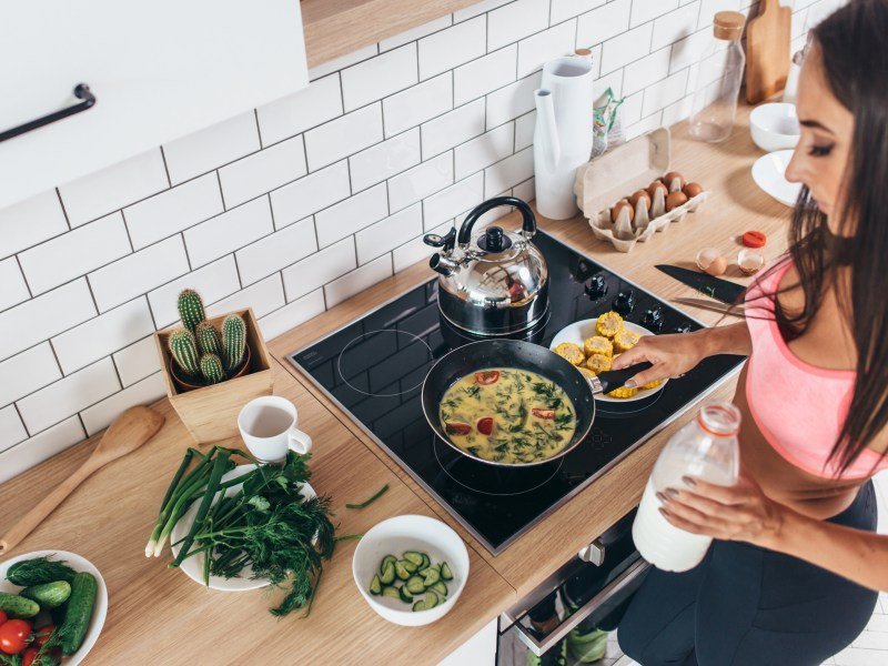 Woman cooking an omelet and holding milk in her left hand. This article discusses if it's safe to do keto while breastfeeding.