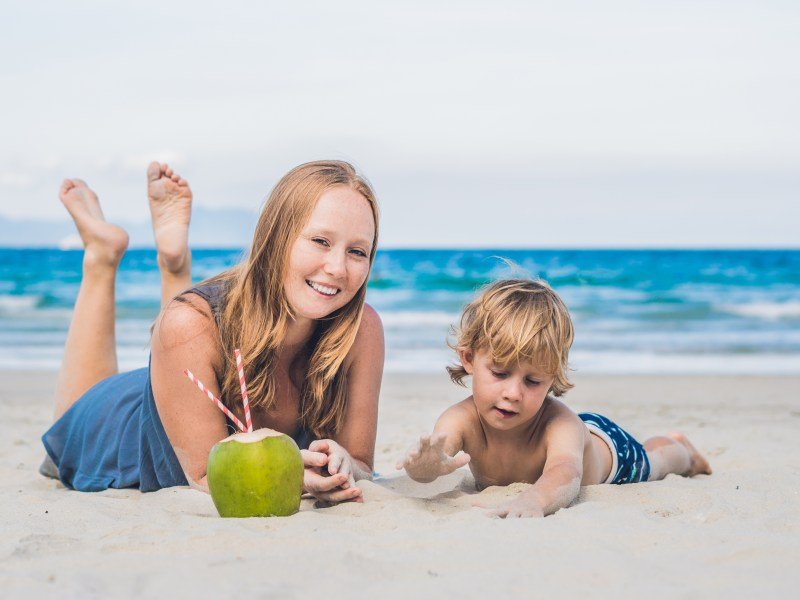 Mom and child laying on stomachs on beach with a coconut in front of them. This articles asks: Does coconut water help with breast milk supply?