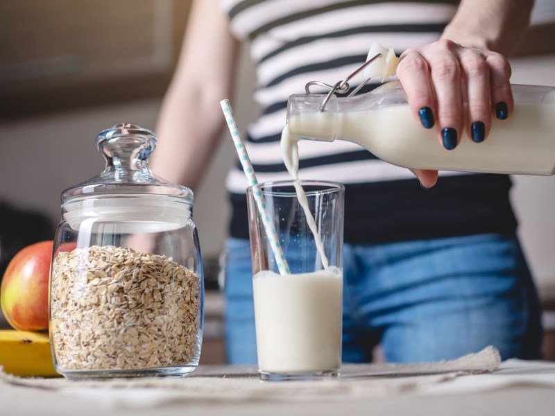 Woman pouring milk into a glass on a kitchen counter with a glass jar of oats next to the glass. This article discusses using oat milk for breast milk supply.