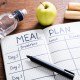 Meal plan journal, pen, bottle or water, and apple on a table. This article reviews breastfeeding meal plan samples.